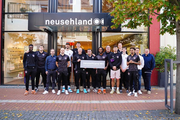 Spieler der GIESSEN 46ers stehen vor dem Neusehland Standort in Gießen und präsentieren ein Neusehland Schild