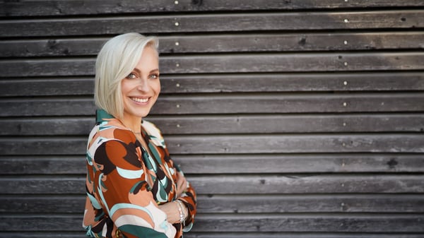 Eine lächelnde Frau mit kurzen, platinblonden Haaren im Profil vor einer grauen Holzwand. Sie trägt eine gemusterte Bluse in Orange, Braun und Grün und blickt direkt in die Kamera.