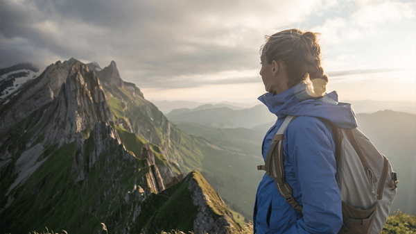 Eine Wanderin mit Rucksack und blauer Outdoor-Jacke blickt von der Seite auf eine beeindruckende Gebirgskette mit steilen, grünen Berghängen im sanften Licht des Spätnachmittags.