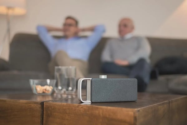 Ein Sprachverstärker liegt auf einem Couchtisch, daneben stehen zwei Wassergläser und ein kleines Schälchen mit Knabberkram, im Hintergrund sitzen zwei Personen und schauen TV