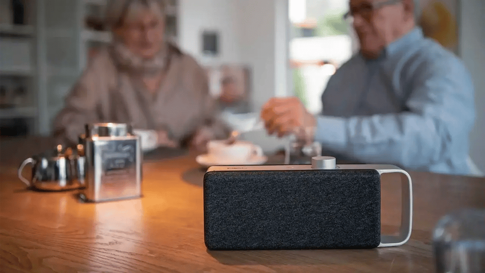 Ein moderner, tragbarer Lautsprecher mit dunkelgrauem Stoffbezug und silbernem Tragegriff steht im Fokus auf einem Holztisch. Im unscharfen Hintergrund sitzt ein Paar beim Kaffeetrinken.