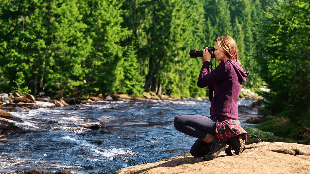 Eine Frau in Wanderbekleidung hockt am felsigen Ufer eines wilden Flusses und fotografiert die Landschaft mit einer Kamera. Im Hintergrund erstreckt sich ein dichter, grüner Nadelwald.