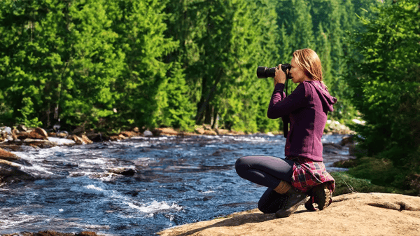 Eine Frau in Wanderbekleidung hockt am felsigen Ufer eines wilden Flusses und fotografiert die Landschaft mit einer Kamera. Im Hintergrund erstreckt sich ein dichter, grüner Nadelwald.