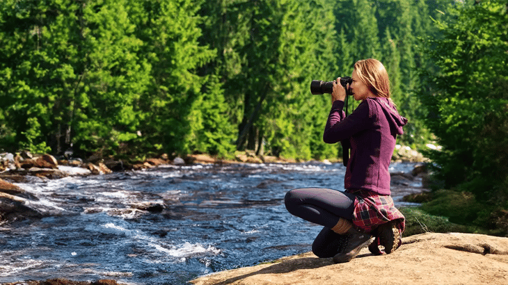 Eine Frau in Wanderbekleidung hockt am felsigen Ufer eines wilden Flusses und fotografiert die Landschaft mit einer Kamera. Im Hintergrund erstreckt sich ein dichter, grüner Nadelwald.