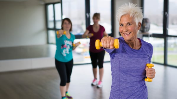 Eine lächelnde Seniorin mit kurzen weißen Haaren macht Sport in einem Fitnessstudio. Sie trägt ein lila Shirt und hockt leicht, während sie eine gelbe Hantel mit dem linken Arm nach vorne boxt. Im unscharfen Hintergrund sind zwei weitere Frauen beim Training zu sehen.