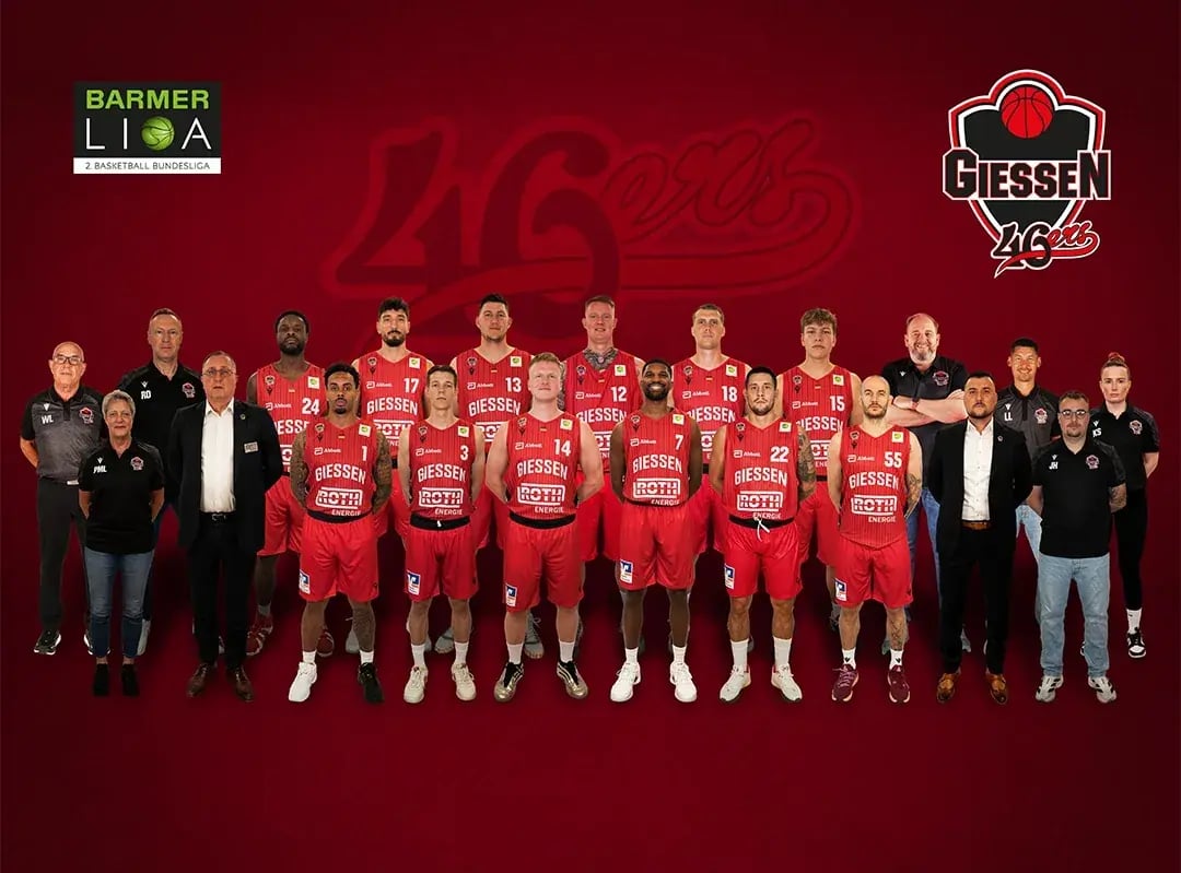 Mannschaftsfoto Giessen 46ers: Spieler in roten Trikots und Trainerstab vor dunkelrotem Hintergrund.