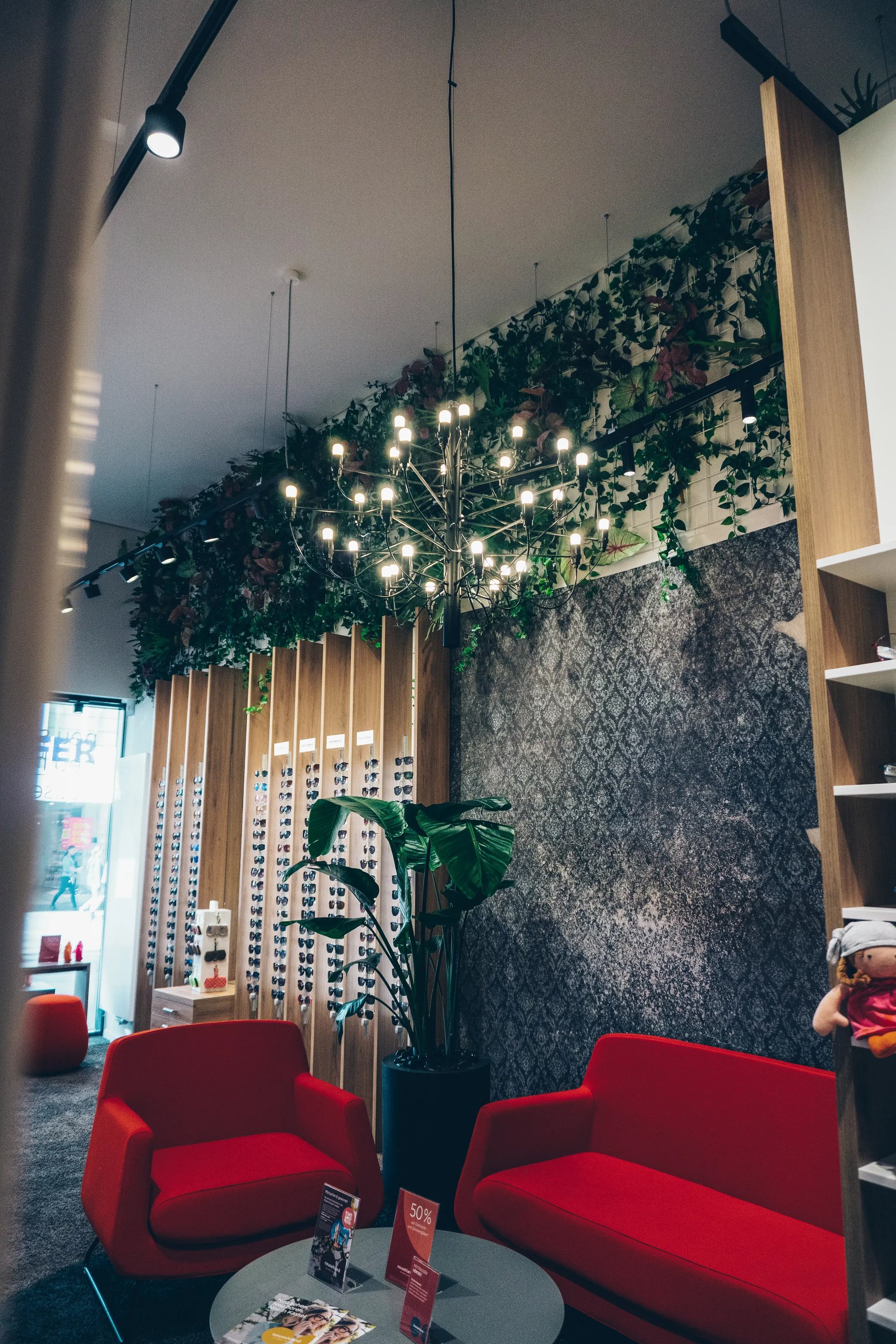 Optikergeschäft mit roten Sofas, Kronleuchter, grüner Pflanzenwand und Brillenregal