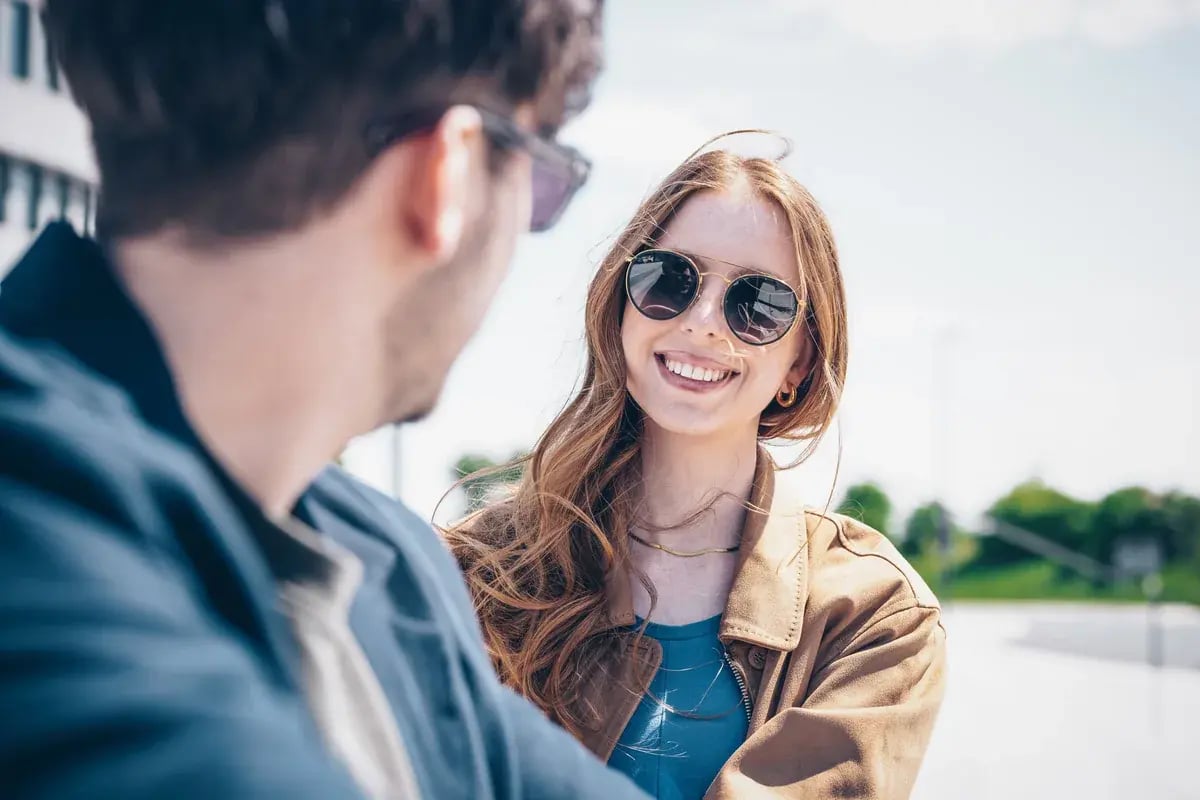 Junge Frau mit runder Sonnenbrille lächelt beim Gespräch im Sonnenlicht