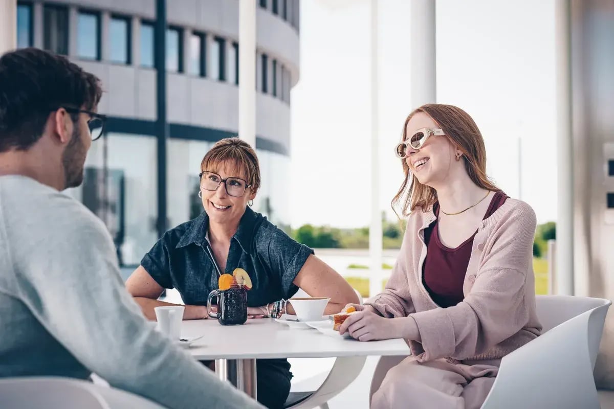 Drei Personen sitzen an einem Tisch. Eine Frau lacht und trägt eine Sonnenbrille
