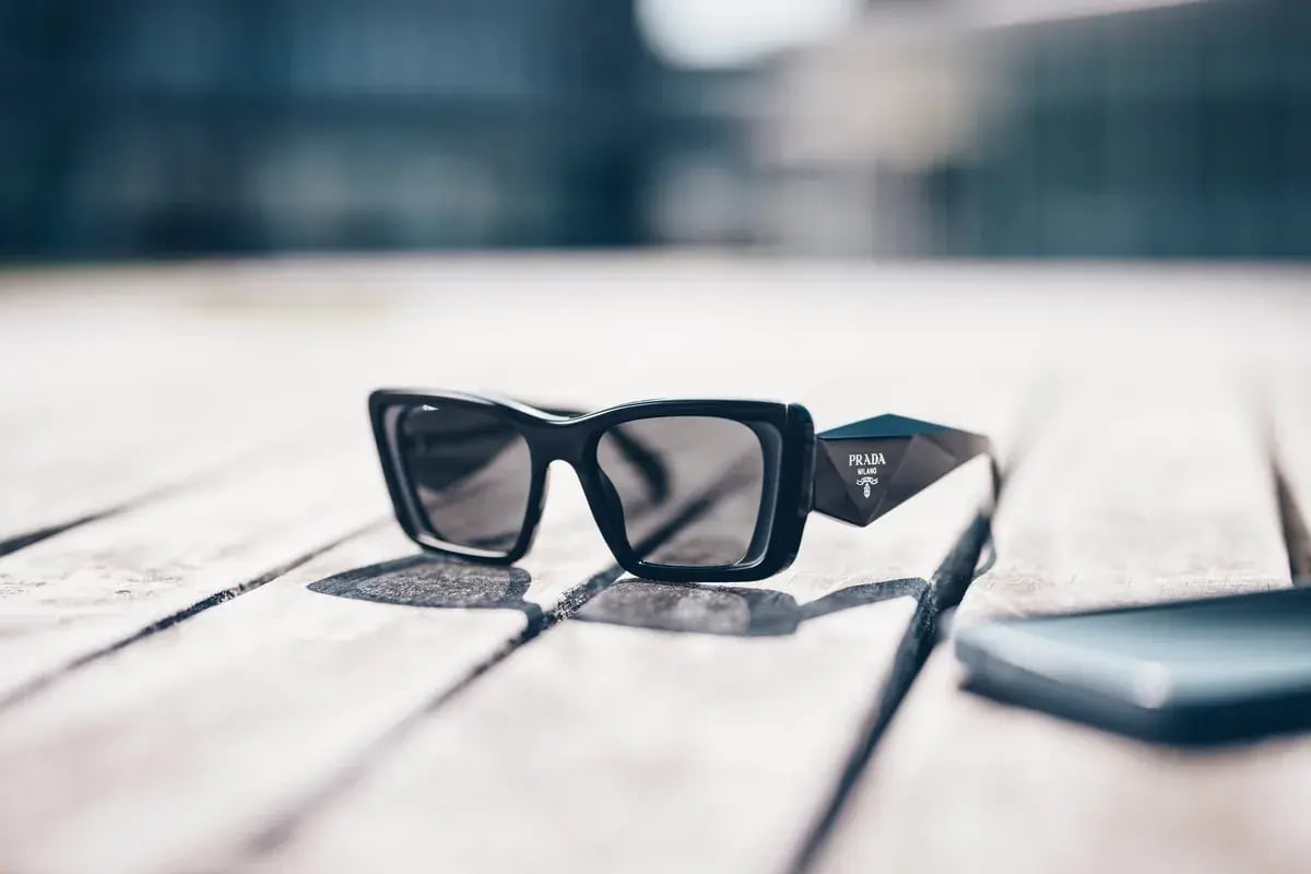 Schwarze Prada-Sonnenbrille mit Dreieck-Logo liegt auf Holzlatten neben Smartphone