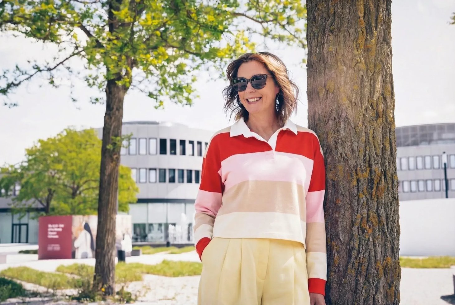 Frau mit Sonnenbrille lehnt an Baumstamm, trägt gestreiften Polo-Pullover und weite Hose, modernes Gebäude im Hintergrund.