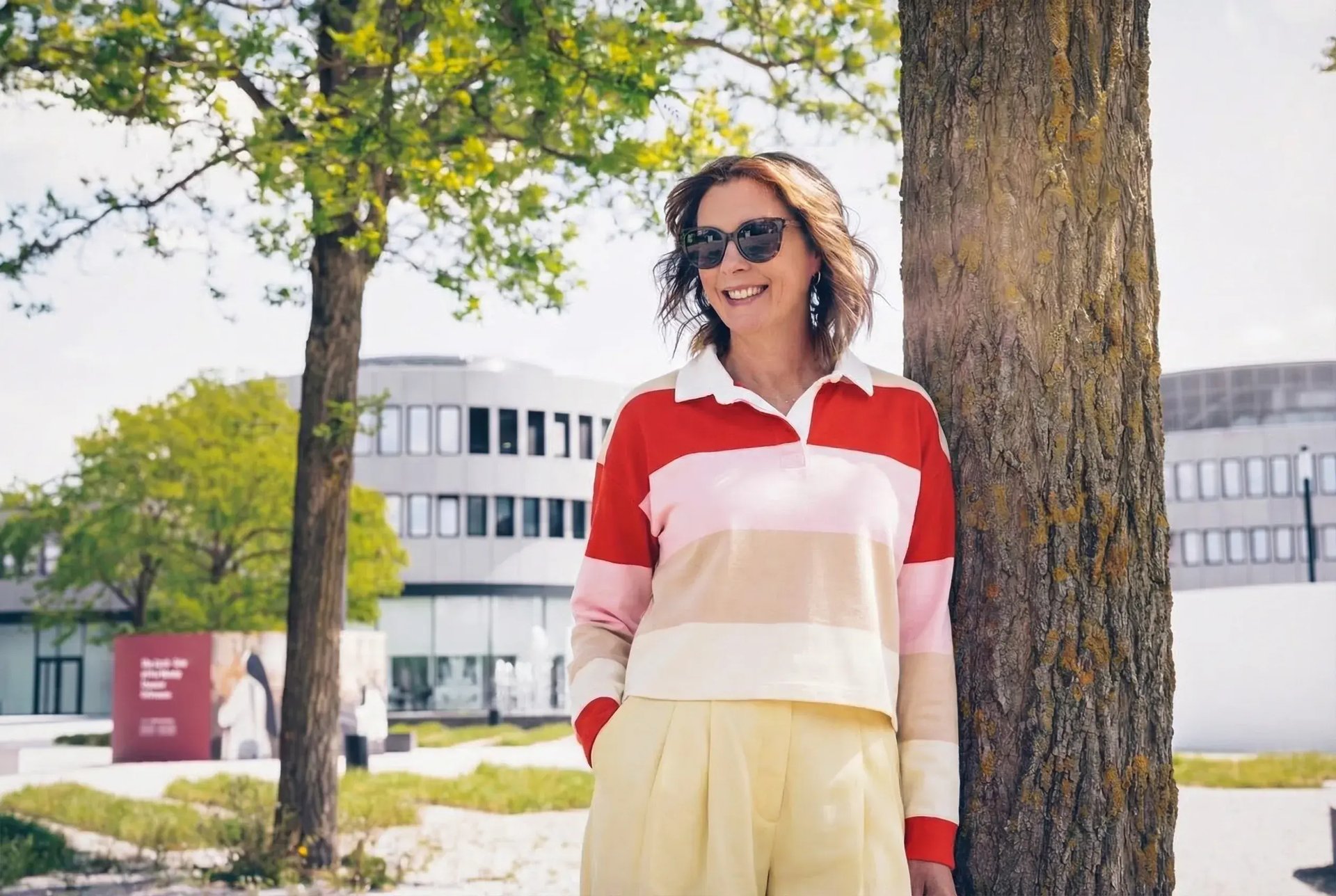 Frau mit Sonnenbrille lehnt an Baumstamm, trägt gestreiften Polo-Pullover und weite Hose, modernes Gebäude im Hintergrund.