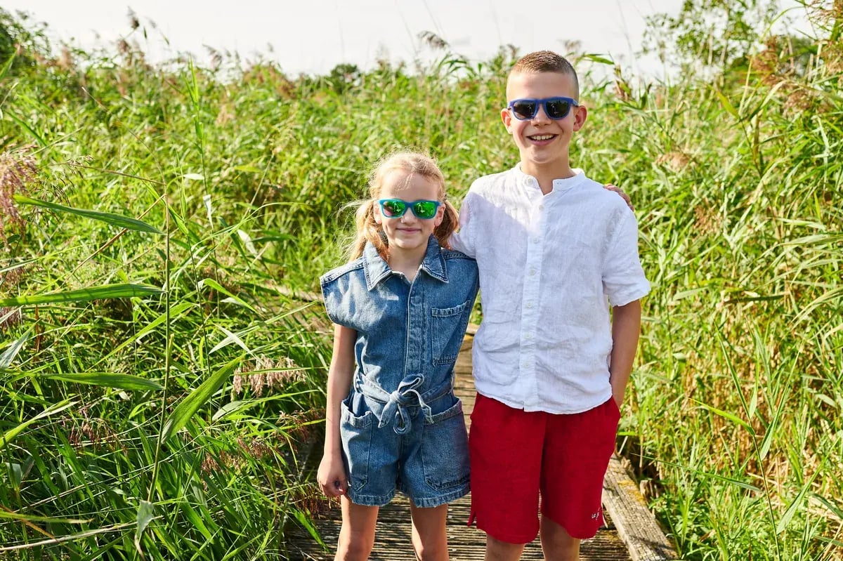 Zwei Kinder mit bunten Sonnenbrillen stehen auf einem Holzsteg im Schilf