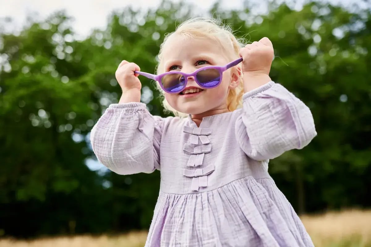 Kleinkind im lila Kleid hält lachend eine lila Sonnenbrille hoch