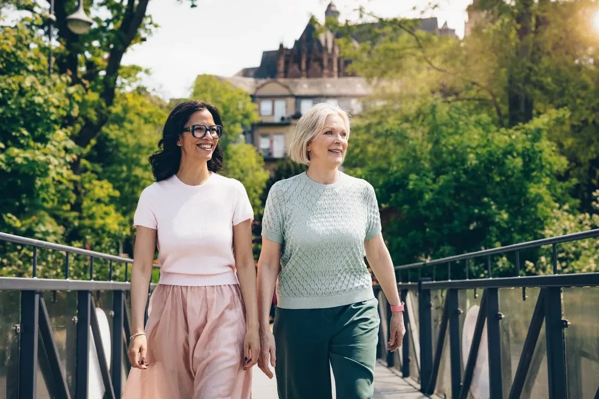 Zwei Frauen spazieren lächelnd auf einer Brücke in einem Stadtpark