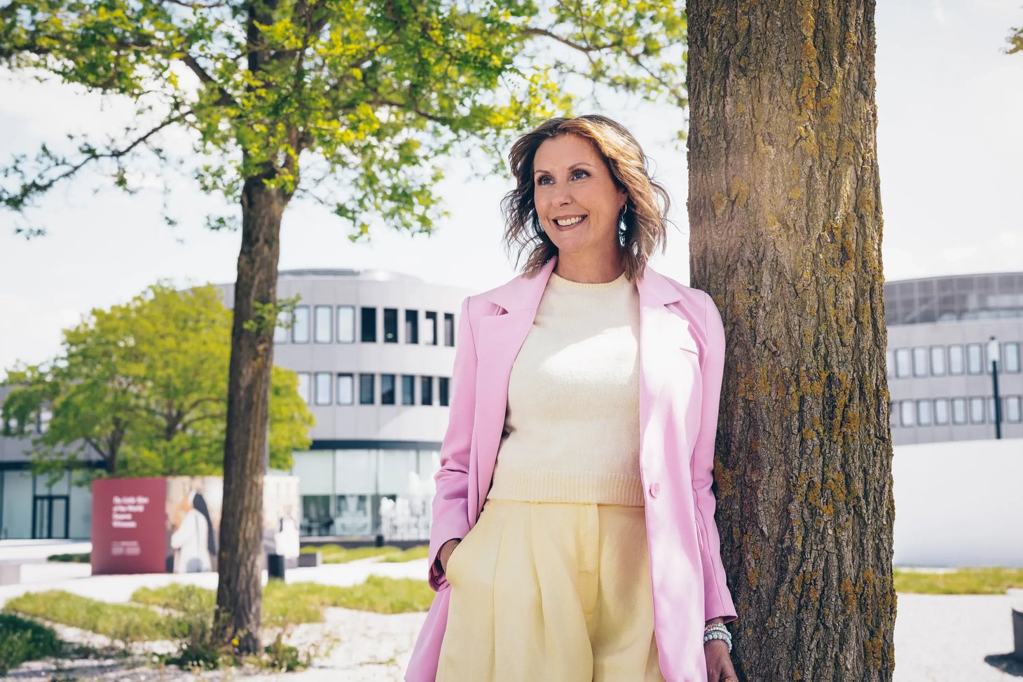 Frau im rosa Mantel steht lächelnd an einem Baum im Freien