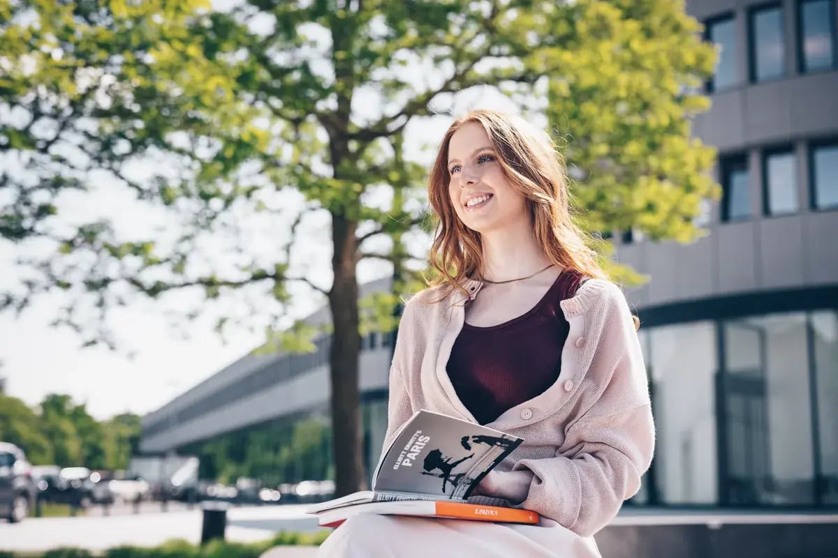 Junge Frau sitzt mit Büchern vor einem Universitätsgebäude im Freien.