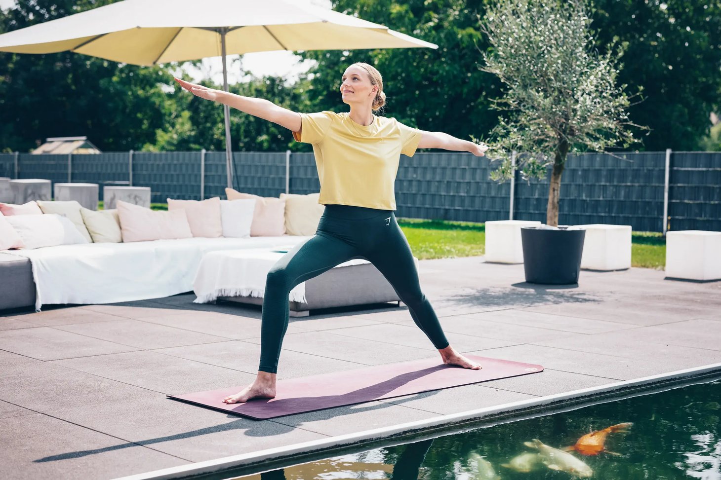 Frau in Krieger-Yoga-Pose auf Matte neben einem Pool mit Koi-Fischen