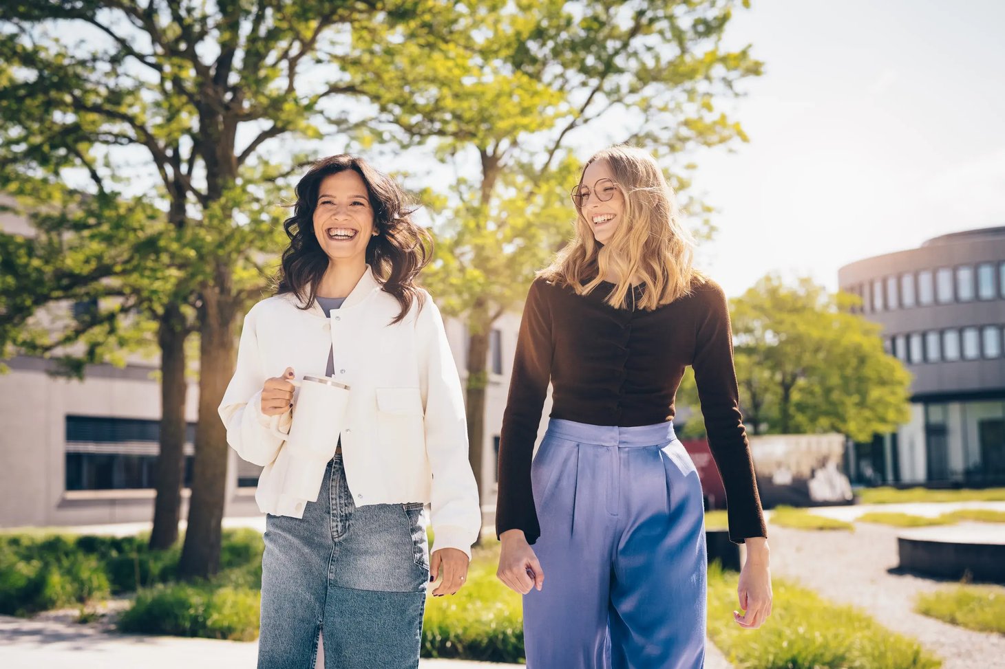 Zwei Frauen gehen nebeneinander auf einem Campus-Außengelände, Bäume und Gebäude im Hintergrund