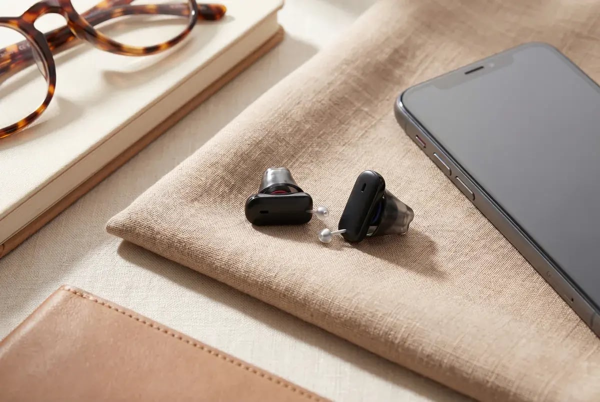 Zwei schwarze Im-Ohr-Hörgeräte auf einem Leinenstoff neben Brille und Smartphone
