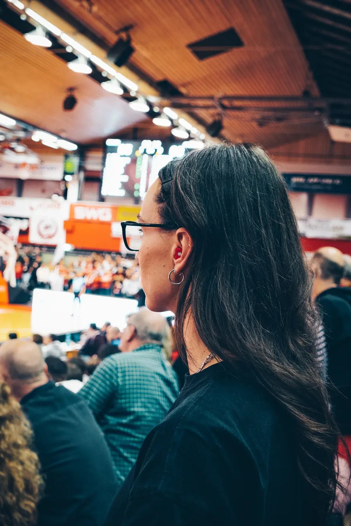 Frau mit rotem Gehörschutz und Brille verfolgt ein Spiel in einer Sporthalle.