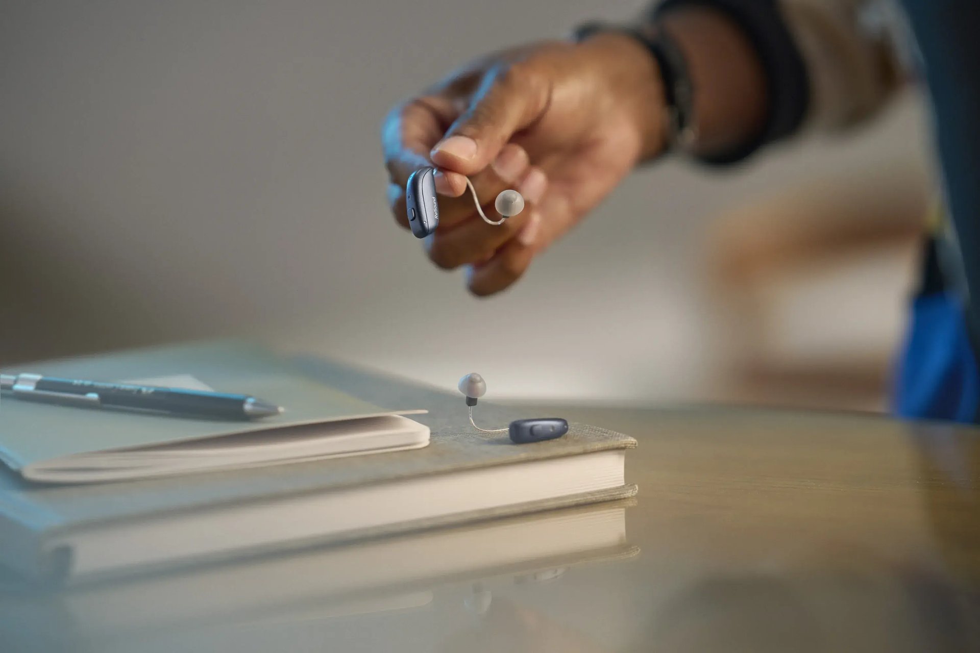 Hand hält ein graues Hörgerät über ein zweites Hörgerät auf einem Bücherstapel