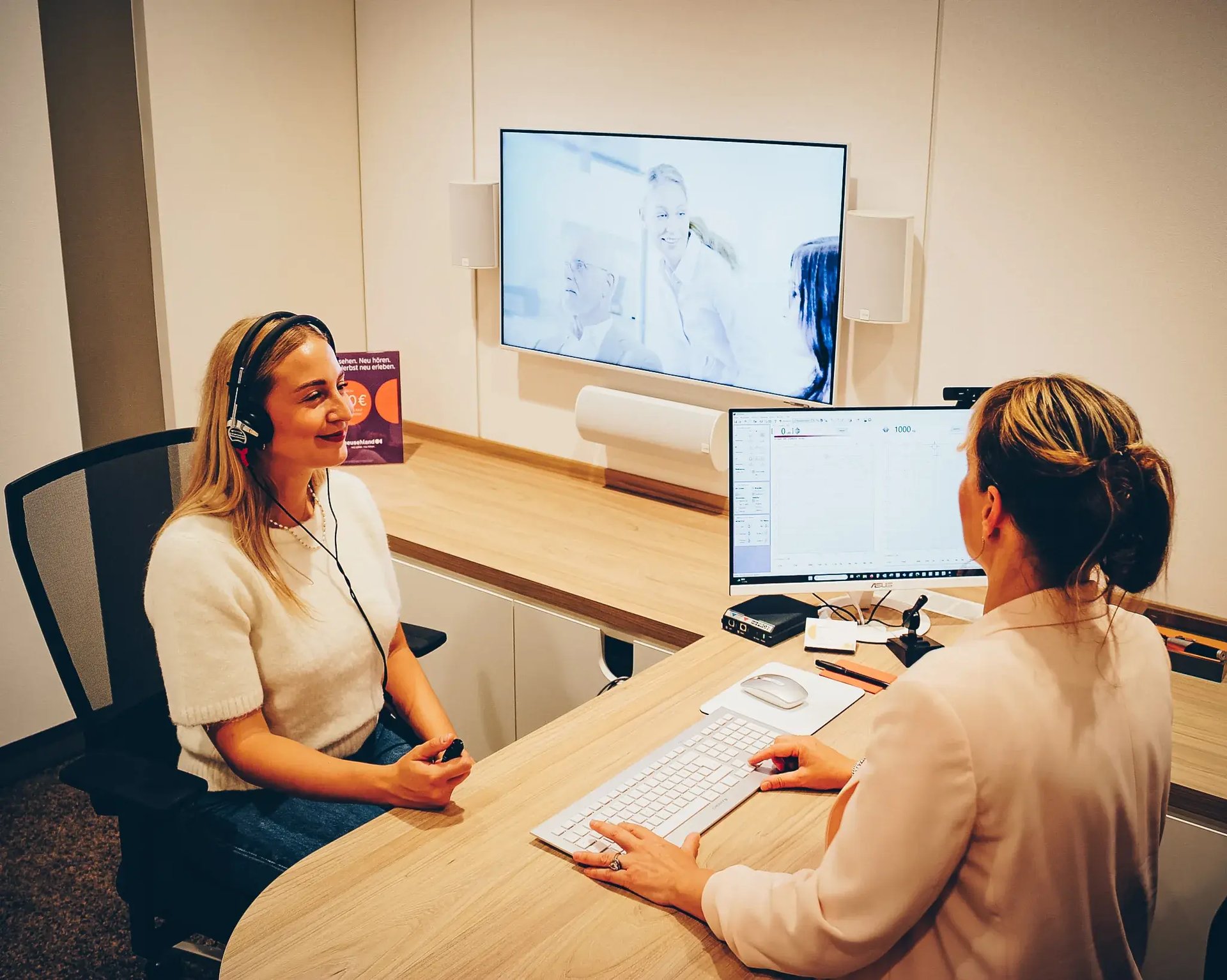 Frau mit Audiometrie-Kopfhörer sitzt einer Beraterin am Tisch gegenüber, PC-Monitor im Vordergrund.