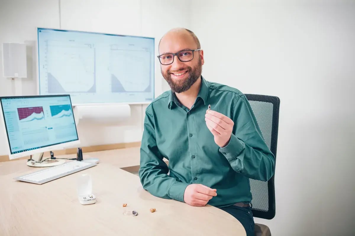 Akustiker hält ein Hörgerät hoch, mehrere Hörgeräte auf dem Schreibtisch