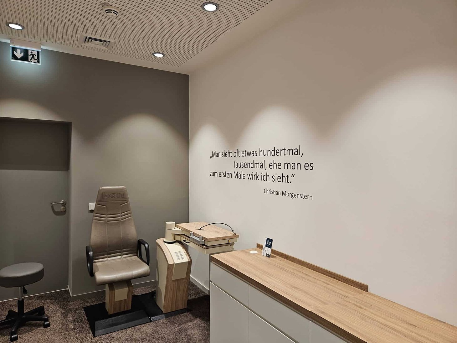 Untersuchungsraum mit Spaltlampe, Patientenstuhl und Zitat von Christian Morgenstern an der Wand