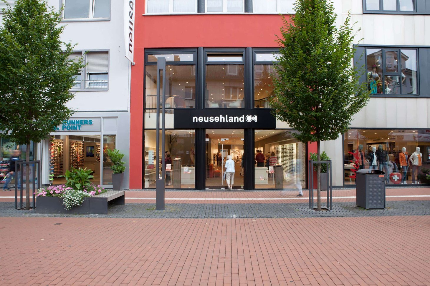Filiale neusehland in Fußgängerzone mit Glasfront, schwarzem Schild und zwei Bäumen