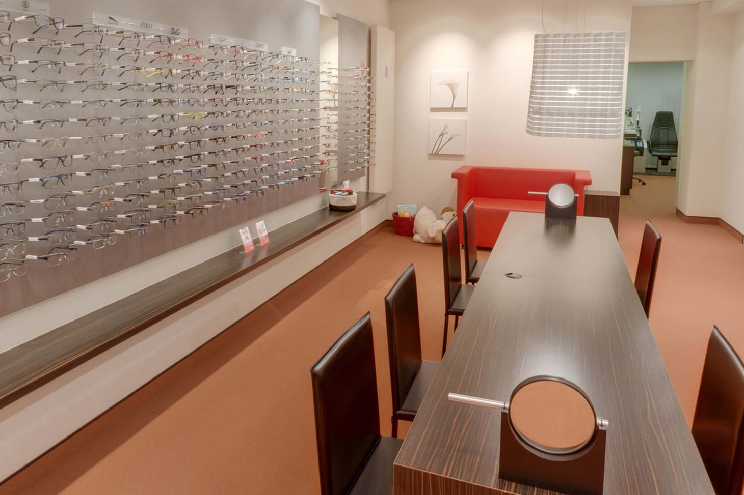 Optiker-Innenraum mit langer Brillenwand links und dunklem Beratungstisch mit Stühlen