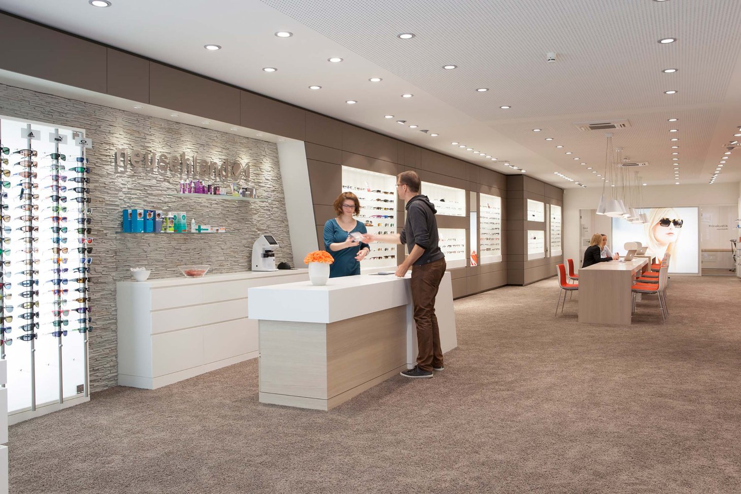 Zwei Personen am Empfangstresen eines Optikers mit Brillenwand im Hintergrund