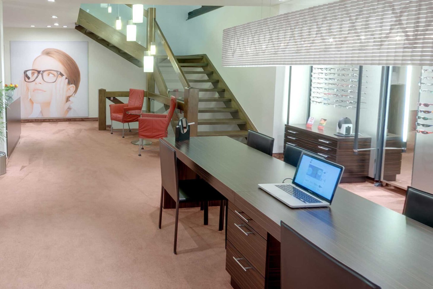 Optiker-Innenraum mit Holztreppe, Beratungstisch mit Laptop und großem Wandbild