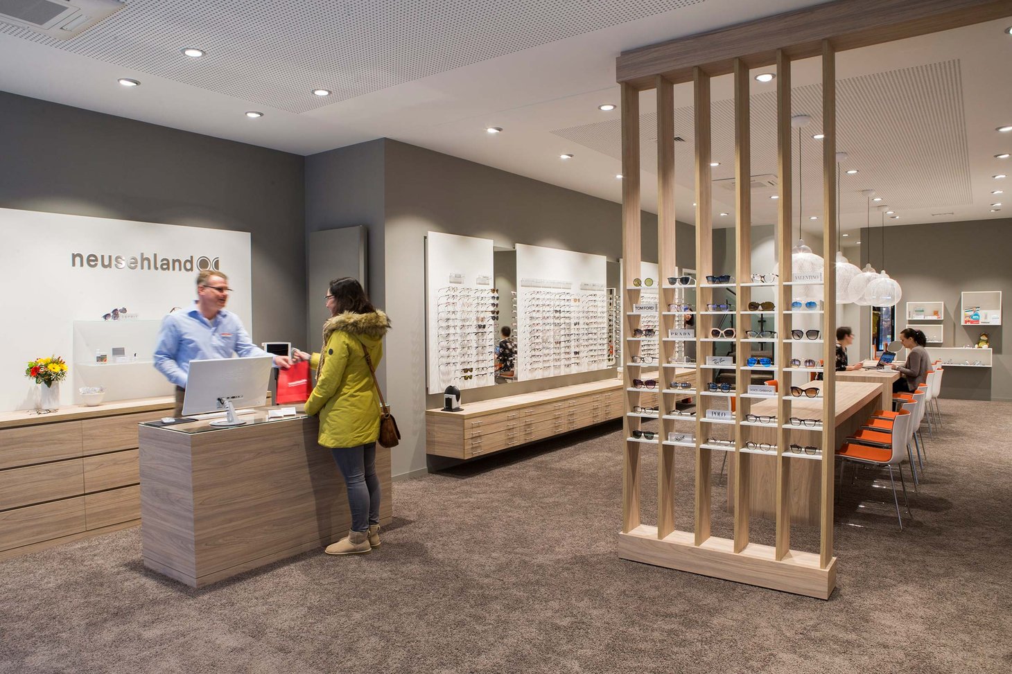 Zwei Personen am Empfangstresen eines Optikers mit Brillenwand im Hintergrund