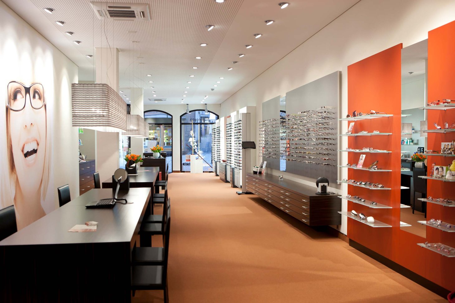 Langer Optikerraum mit orange-weißen Brillenregalen beidseitig und einem Wandbild mit Porträt