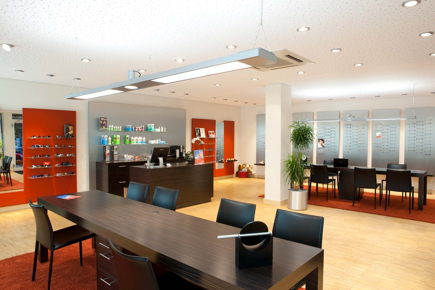 Optikerraum mit dunklem Empfangstresen, orangen Wandakzenten, Brillenregalen und Beratungstischen