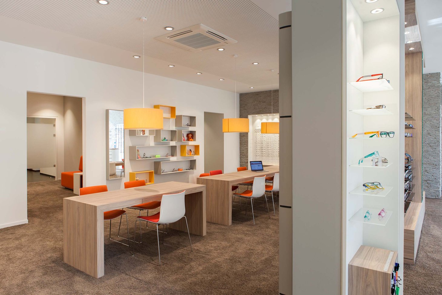 Heller Optikerverkaufsraum mit Holzberatungstischen, orangen Hängelampen und Brillenregal
