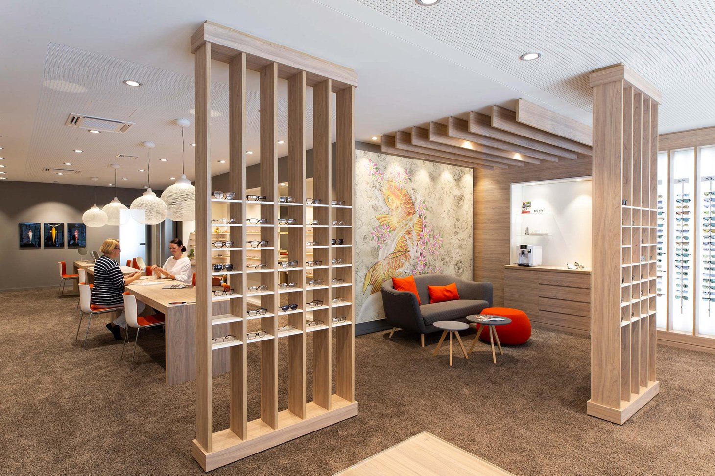 Zwei Personen sitzen an einem Beratungstisch in einem Optikergeschäft mit Holzstelen und Brillenregal
