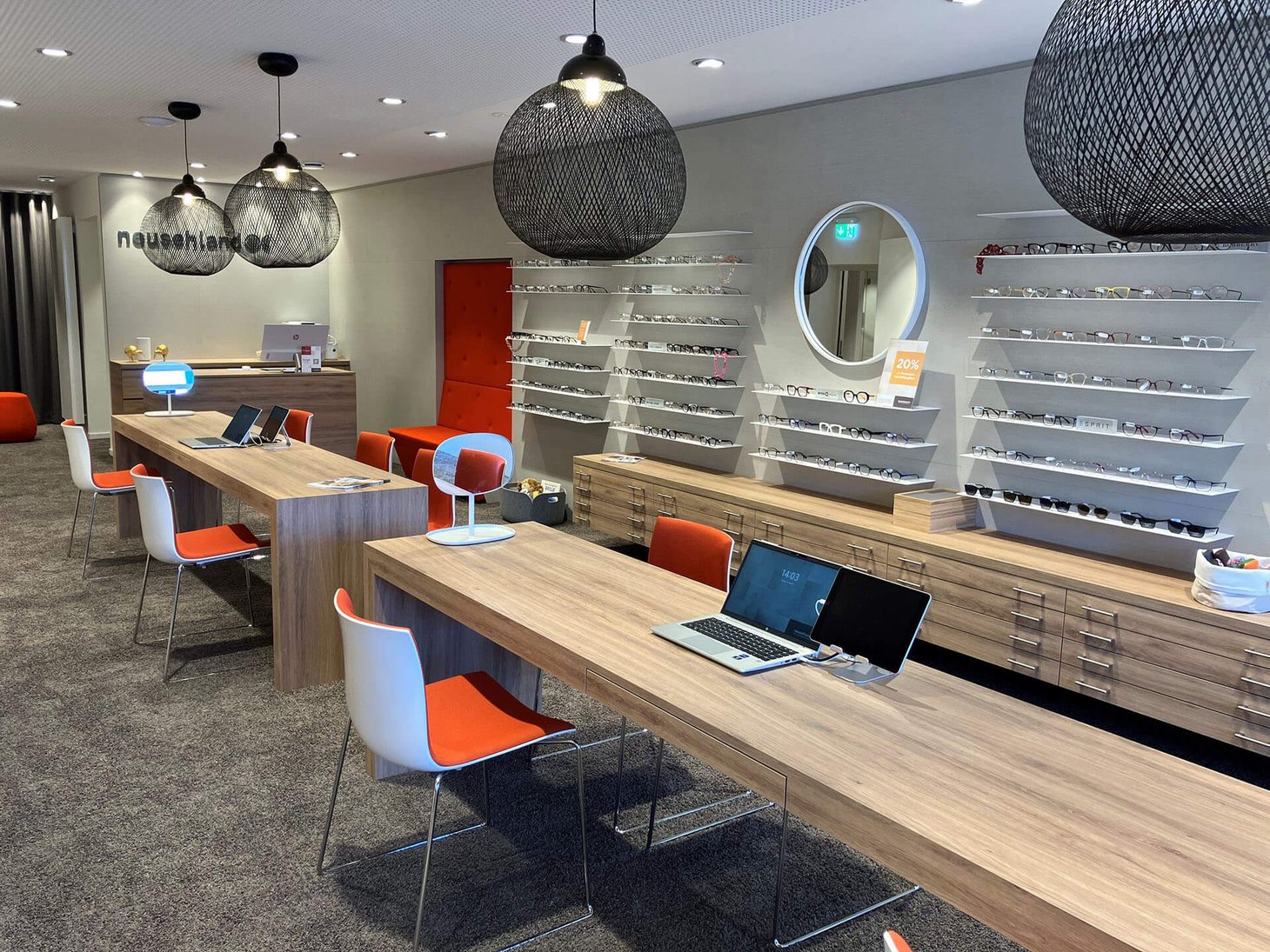 Optikerverkaufsraum mit langen Holztischen, orangefarbenen Stühlen, Hängeleuchten und Brillenwand