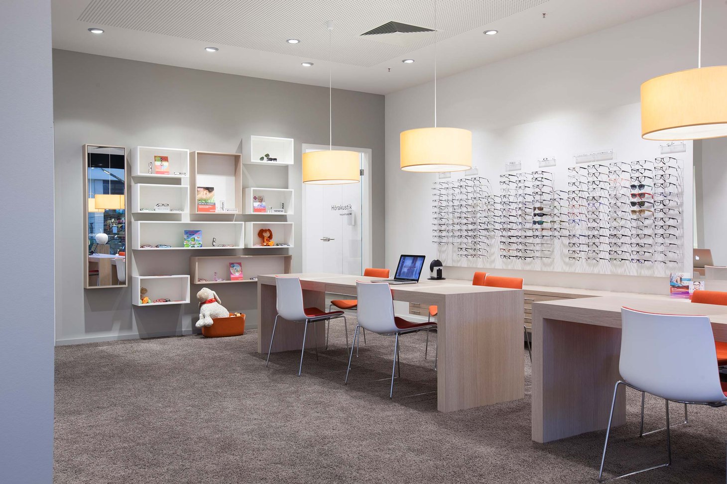 Heller Optiker-Verkaufsraum mit Brillenwand, Beratungstisch und orangefarbenen Stühlen
