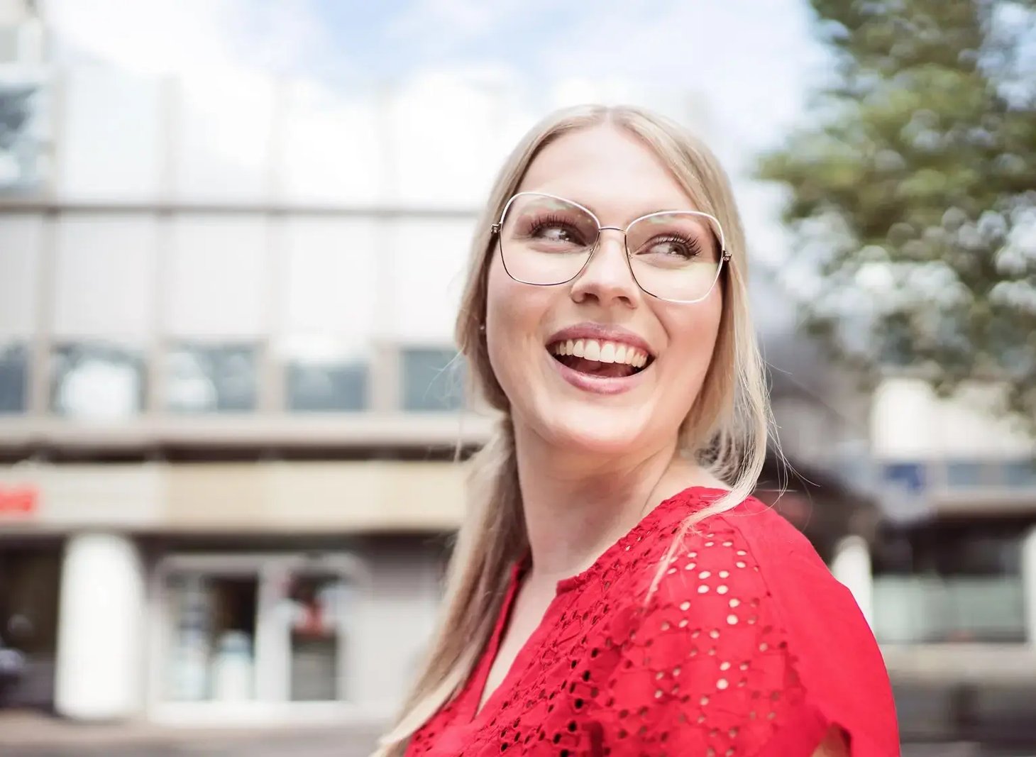Frau mit eckiger Metallbrille und rotem Oberteil lacht, unscharfer Stadtbezirk im Hintergrund