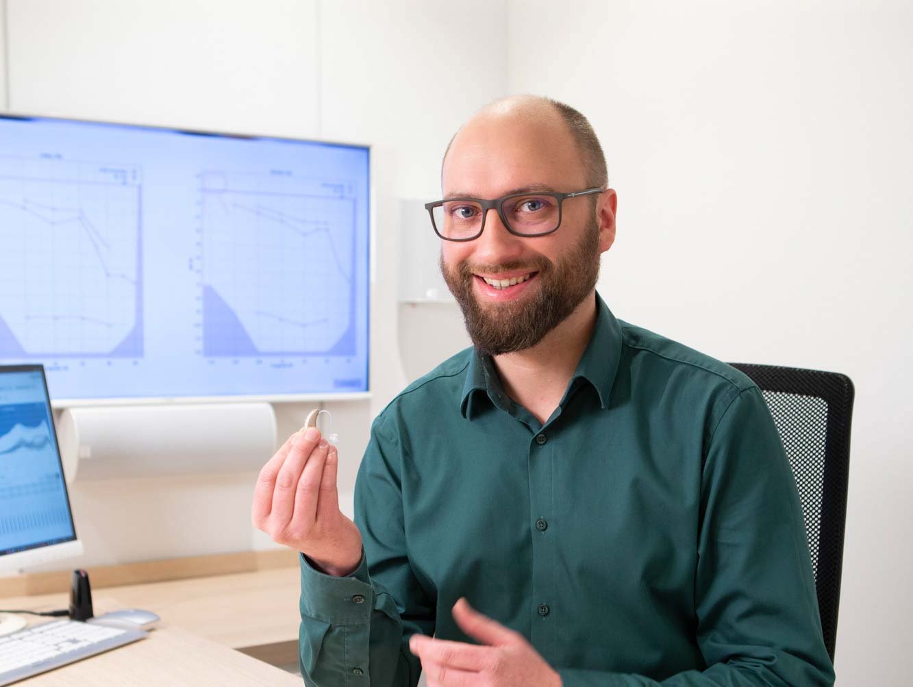 Mann mit Brille sitzt am Schreibtisch und hält ein Hörgerät in der Hand, Audiogramm im Hintergrund