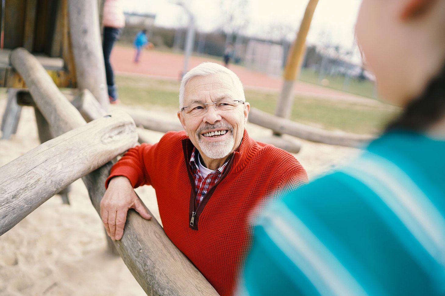 Älterer Mann mit Brille lehnt an einem Klettergerüst auf einem Spielplatz und blickt ein Kind an