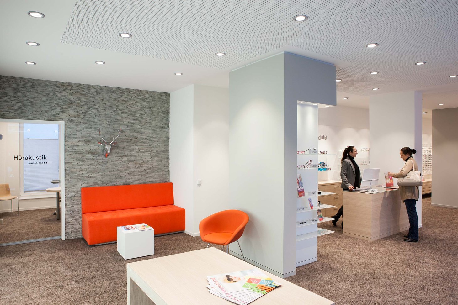 Orangefarbenes Sofa und Sessel vor grauer Steinwand, Schriftzug "Hörakustik"