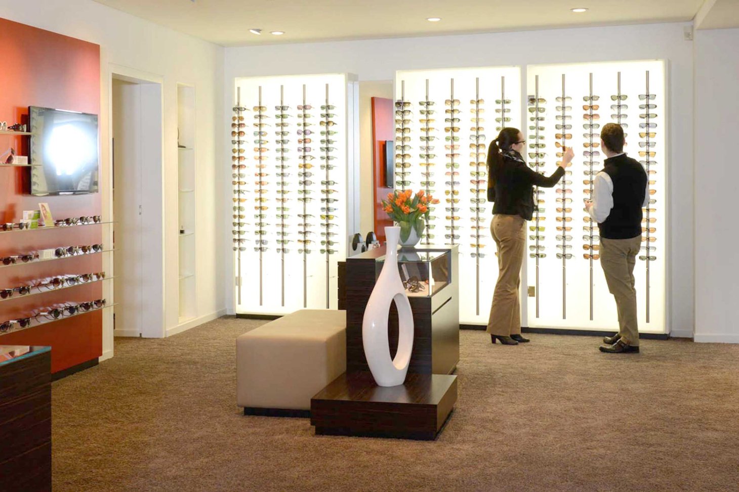 Zwei Personen stehen in einem Optiker-Geschäft vor einer Brillenwand mit zahlreichen Modellen