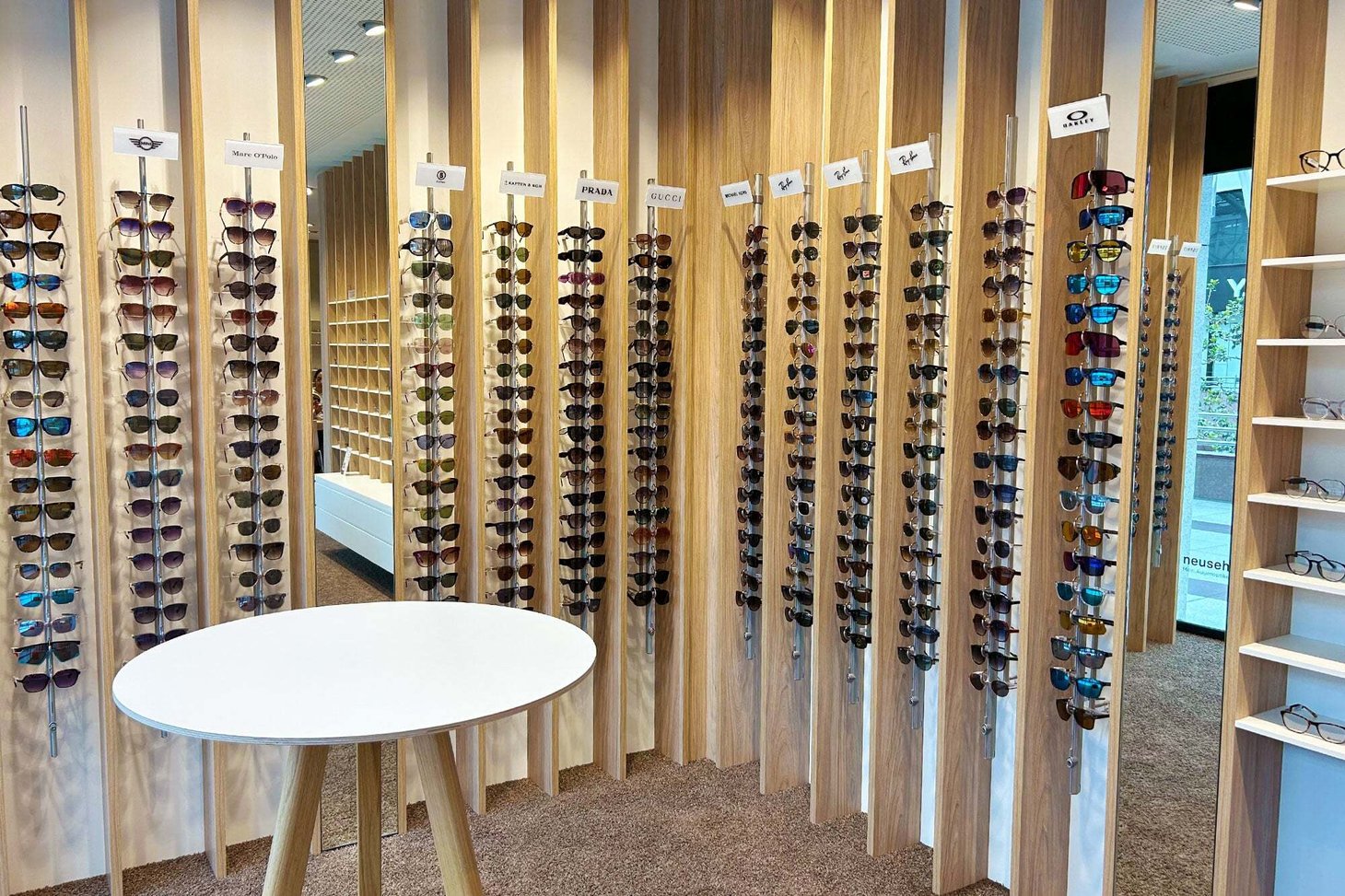 Zahlreiche Sonnenbrillen an Holzstäben-Display mit Markenschildern an der Wand