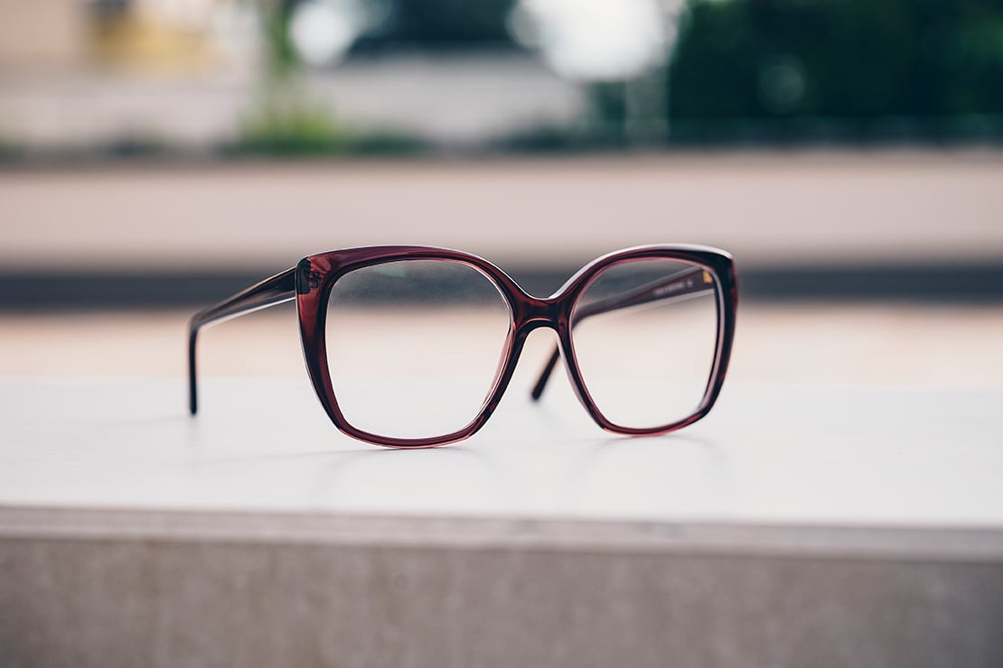 Moderne Damenbrille mit bordeauxrotem Kunststoffrahmen und großen, leicht eckigen Gläsern auf einer hellen Oberfläche.