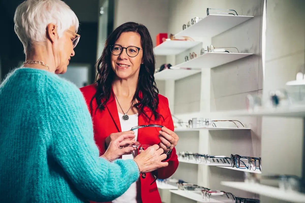 Optikerin im roten Blazer zeigt älterer Kundin Brillengestelle in einem Optikgeschäft.