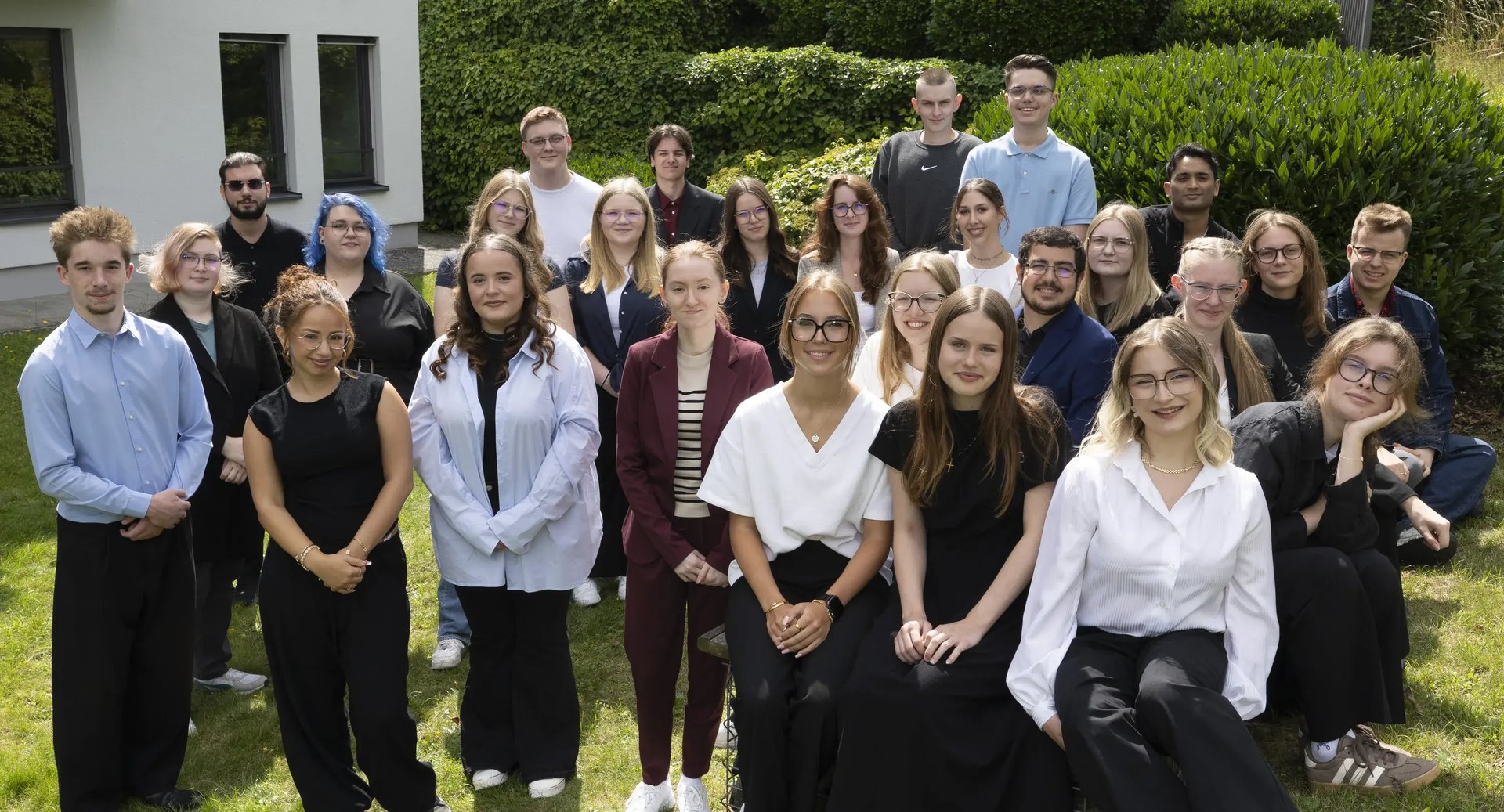 Gruppe von etwa 25 jungen Menschen steht in Reihen auf einer Rasenfläche vor einem Gebäude.