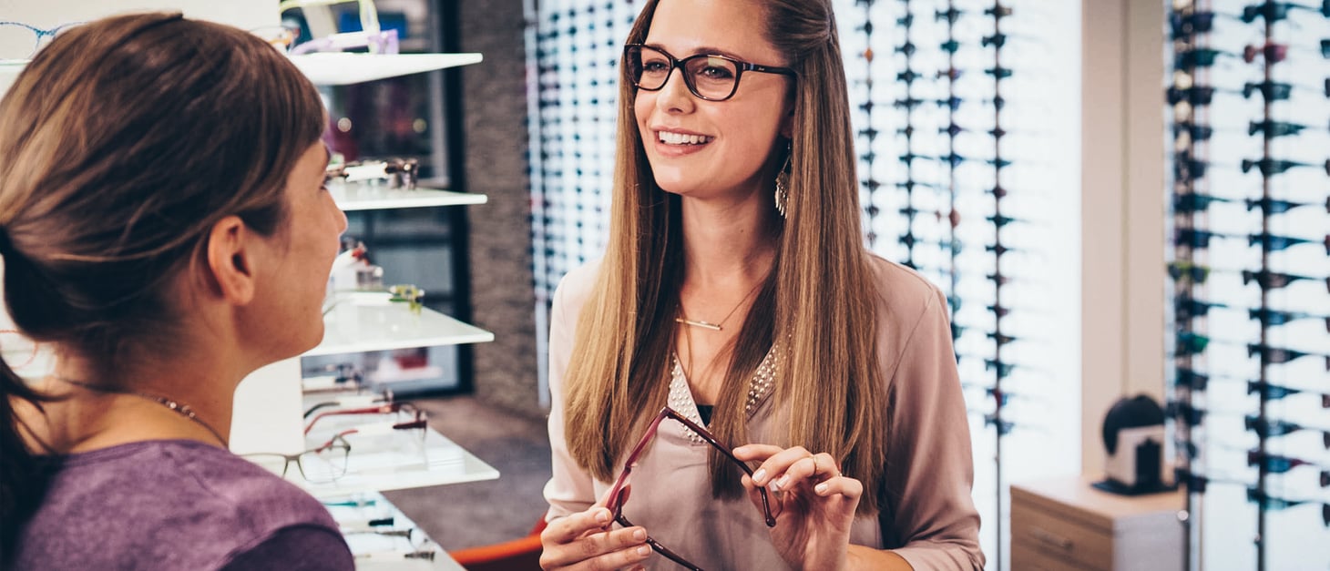 Eine lächelnde Frau mit Brille hält ein Brillengestell in den Händen und lässt sich in einem Optikergeschäft beraten.